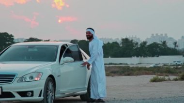 Ulusal Arap kıyafetleri içinde sakallı genç bir işadamı lüks bir arabanın kapısını kapatıyor. Arka planda Sunset ve Dubai gökdelenleri