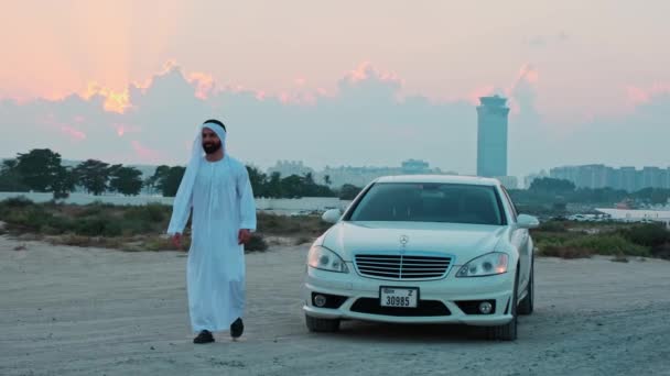 国のアラブの服の若いひげ付きビジネスマンは高価なプレミアムカーの隣で夕日を踊ります ドバイの高層ビルを背景に — ストック動画