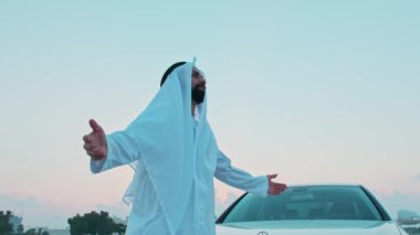 Ulusal Arap elbiseli genç sakallı bir işadamı ellerini pahalı bir arabanın yanına uzatıyor. Arka planda Dubai 'nin günbatımı ve gökdelenleri.