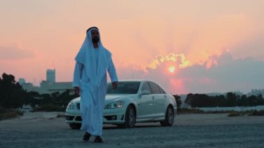 Ulusal Arap elbiseli genç sakallı bir işadamı pahalı arabasından kameraya doğru yürür. Arka planda gün batımı bulutlu gökyüzü.