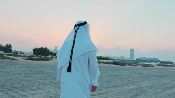 一位身穿阿拉伯民族服装 留着胡子的年轻商人走向他那辆昂贵的汽车 在迪拜摩天大楼的背景下 夕阳西下 — 图库视频影像