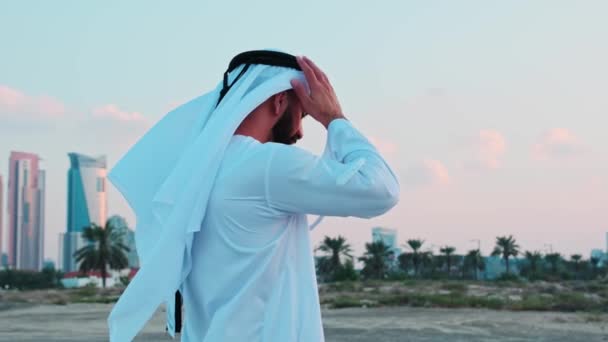 迪拜的摩天大楼前 站着一位身穿阿拉伯民族服装 留着胡子的年轻商人 在多云的天空中落日 — 图库视频影像