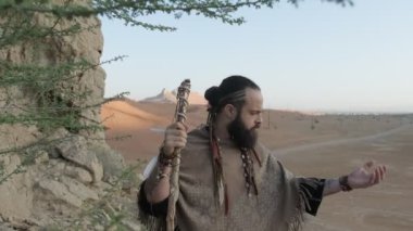 Elinde asası olan genç sakallı bir adam çölün kumlu kumları arasında bir kayanın üzerinde dua ediyor. Ağır çekim, BAE