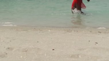 Bikinili esmer ve açık kırmızı elbiseli genç bir kadın sörf suyunda kumlu bir sahilde yalınayak yürüyor. Yavaş çekim
