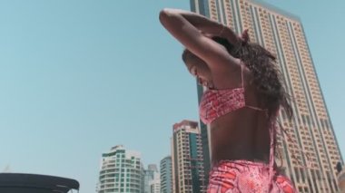 Dubai gökdelenlerinin arka planına karşı özel bir teknenin güvertesinde kısa parlak elbiseli esmer bir kadın. BAE, Ağır çekim