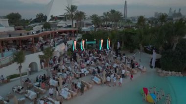 Bir İHA açık havuzun üzerinde, deniz kenarında bir sopayla uçuyor, Dubai. İnsanlar güneşleniyor, barda ve kafede dinleniyor, havuzda yüzüyor. Hava Görünümü.