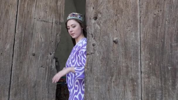 一个年轻的女人 穿着白色的短裙 身穿东方长袍 背靠木门 乌兹别克斯坦慢动作 — 图库视频影像