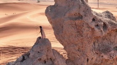 Bir dron, Birleşik Arap Emirlikleri 'nin kumlu çölündeki bir uçurumda dua eden şaman kılığında sakallı bir adamın etrafında uçuyor. Hava görünümü
