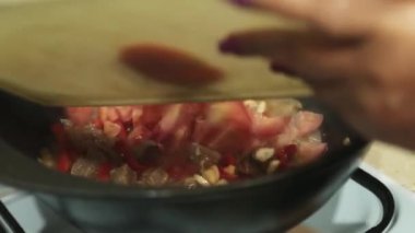 Yaşlı bir kadın, doğranmış domatesleri kızgın yağlı bir kazana koyar ve ahşap bir kaşıkla karıştırır. Mutfak, gaz ocağı, yakın çekim..
