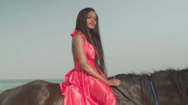 Kırmızı elbiseli genç esmer bir kadın okyanus kıyısında duran bir atın üzerinde oturuyor. Yavaş çekim.