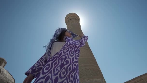 一位身穿白衣和东方长袍的年轻女子在一座古老的尖塔旁边的阳光下跳舞 乌兹别克斯坦慢动作 — 图库视频影像