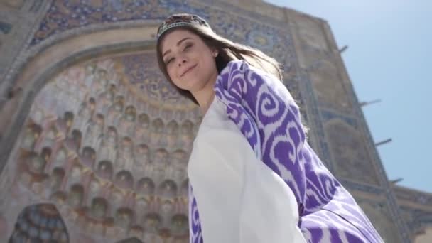 一位身穿东方长袍头戴便帽的年轻女子站在一座古寺附近的风中 乌兹别克斯坦慢动作 — 图库视频影像