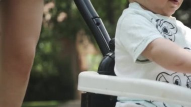 Şortlu genç bir anne, küçük oğlunu oyuncak bir arabaya bindiriyor. Park, güneşli bir gün. Yavaş Hareket, Kapat.