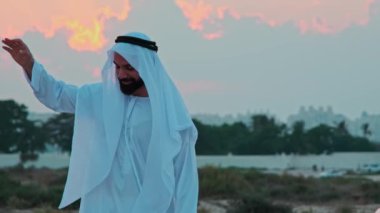 Ulusal Arap kıyafetleri içinde sakallı genç bir işadamı gün batımının arka planında dans ediyor. Dubais gökdelenleri ufukta görünüyor..