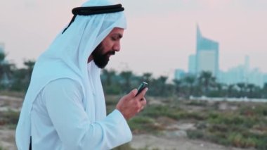Ulusal Arap kıyafetleri içinde sakallı genç bir işadamı cep telefonuna bakıyor. Arkadaki Dubai gökdelenlerinden sipariş verin..
