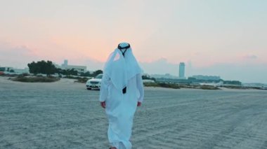 Ulusal Arap elbiseli genç sakallı bir işadamı pahalı arabasına doğru yürür. Dubai 'nin gökdelenlerinin arkasında gün batımı.