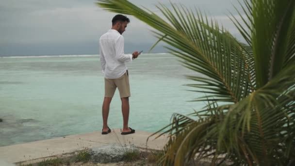 휴대폰을 남자는 야자수 해변에 있습니다 바다의 파도와 하늘의 배경에 대하여 — 비디오