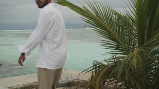 軽い服を着た若者は ヤシの木の下の砂浜に沿って歩いている 海の波と曇りの空の背景について モルディブ スローモーション — ストック動画