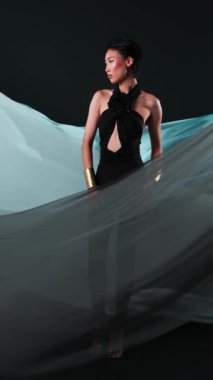 Siyah tulumlu ve parlak makyajlı Asyalı bir gencin dikey stüdyo portresi. Rüzgarda çırpınan bir kumaş