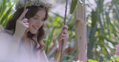 A young beautiful girl in a wreath of flowers swings on a swing among a green garden. Slow motion
