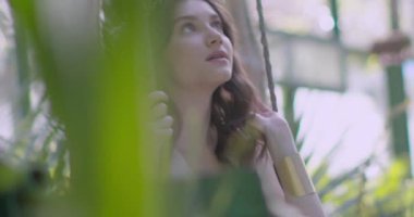 A young beautiful girl in a wreath of flowers swings on a swing among a green garden. Slow motion