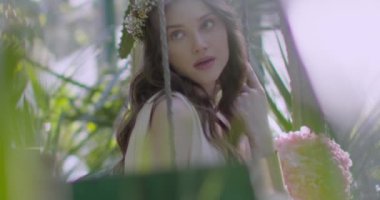 A young beautiful girl in a wreath of flowers swings on a swing among a green garden. Slow motion