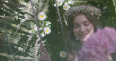 A young beautiful girl in a wreath of flowers swings on a swing among a green garden. Slow motion