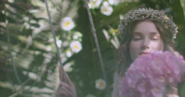 A young beautiful girl in a wreath of flowers swings on a swing among a green garden. Slow motion