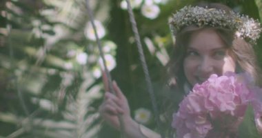 A young beautiful girl in a wreath of flowers swings on a swing among a green garden. Slow motion