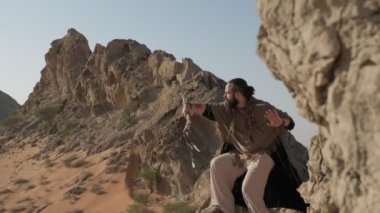 Elinde asası ve şaman kıyafetleri olan bir adam çölün kumları arasında bir kayanın üzerinde duruyor ve dans ediyor. Gökyüzü açık, yavaş çekim