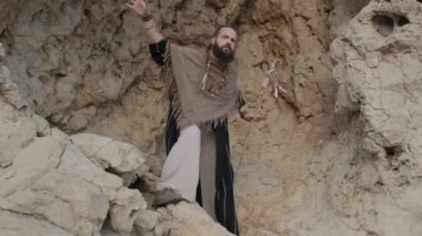 Elinde asası olan genç sakallı bir adam çölün kumlu kumları arasında bir kayanın üzerinde dua ediyor. Ağır çekim, BAE