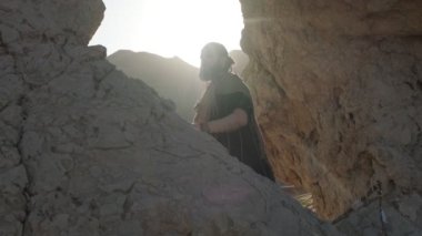 Genç sakallı bir şaman çölün kum tepelerinin arasında bir kaya mağarasında dururken dua eder. Yavaş çekim