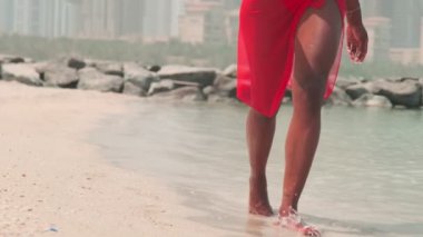 Bikinili esmer ve açık kırmızı elbiseli genç bir kadın denizin ve Dubai 'nin gökdelenlerinin önünde çıplak ayakla kayalarda yürüyor. BAE, Ağır çekim.