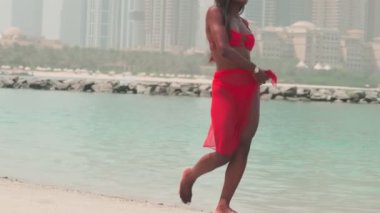 Bikinili esmer ve açık kırmızı elbiseli genç bir kadın denizin ve Dubai 'nin gökdelenlerinin önünde çıplak ayakla kayalarda yürüyor. BAE, Ağır çekim.