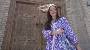 Uzun saçlı, kısa beyaz elbiseli ve ahşap bir kapının arkasında oryantal cüppeli genç bir kadın. Yavaş Hareket, Özbekistan