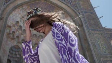Doğulu cüppeli ve şapkalı, uzun saçlı genç bir kadın eski bir caminin yanında rüzgârda duruyor. Yavaş Hareket, Özbekistan
