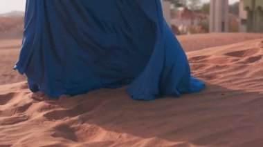Uzun mavi elbiseli dalgalı saçlı genç bir kadın kum kaplı şehrin yanındaki çöl kumullarında yalınayak yürüyor. Yavaş Hareket