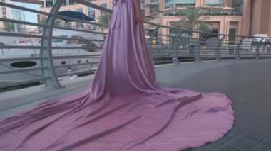 Sarışın ve uzun mor elbiseli genç bir kadın marina boyunca Dubais gökdelenlerinin önünde yürüyor. Yavaş Hareket.