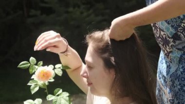 Yetişkin bir anne, genç kızlarının uzun saçlarını gül çalılarının yanındaki meyve bahçesinde şekillendirir. Yavaş Hareket