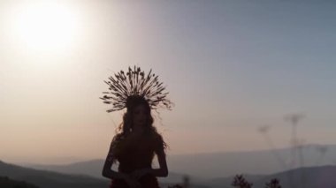Sonbahar giysisi, çiçekli ve otlu bir kadın ve mısır kulaklı bir başlık dağlarda günbatımının arka planında duruyor. Yavaş Hareket.