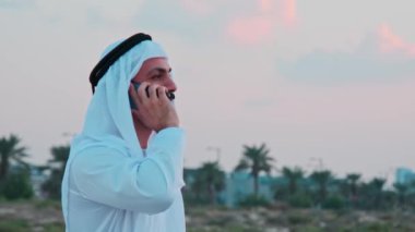 Ulusal Arap kıyafetleriyle telefonda konuşan sakallı bir iş adamının portresi. Arka planda gün batımı bulutlu gökyüzü.