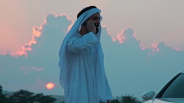 Ulusal Arap kıyafetleri içinde sakallı genç bir işadamı pahalı bir arabanın yanında telefonla konuşuyor. Arka planda gün batımı bulutlu gökyüzü.