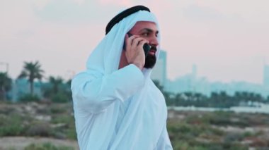 Ulusal Arap kıyafetleri içinde sakallı genç bir işadamı cep telefonuna bakıyor. Arkadaki Dubai gökdelenlerinden sipariş verin..