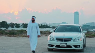 Ulusal Arap kıyafetleri içinde sakallı genç bir işadamı beyaz premium bir arabanın yanında duruyor. Arka planda Dubai 'nin günbatımı ve gökdelenleri.