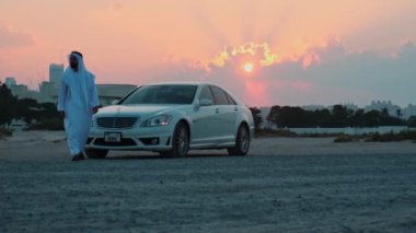 Ulusal Arap elbiseli genç sakallı bir işadamı pahalı arabasından kameraya doğru yürür. Arka planda gün batımı bulutlu gökyüzü.