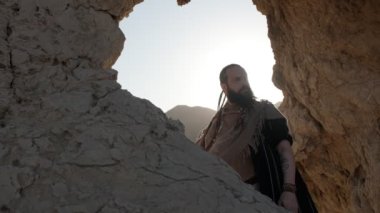 Genç sakallı bir şaman çölün kum tepelerinin arasında bir kaya mağarasında dururken dua eder. Yavaş çekim