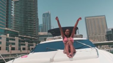 Dubai gökdelenlerinin arka planına karşı özel bir teknenin güvertesinde kısa parlak elbiseli esmer bir kadın. BAE, Ağır çekim