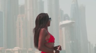 Bikinili esmer ve açık kırmızı elbiseli genç bir kadın denizin ve Dubai 'nin gökdelenlerinin önünde çıplak ayakla kayalarda yürüyor. BAE, Ağır çekim.