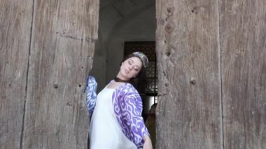 Uzun saçlı, kısa beyaz elbiseli ve ahşap bir kapının arkasında oryantal cüppeli genç bir kadın. Yavaş Hareket, Özbekistan