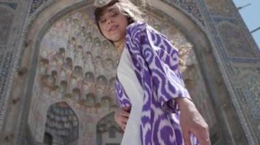 Doğulu cüppeli ve şapkalı, uzun saçlı genç bir kadın eski bir caminin yanında rüzgârda duruyor. Yavaş Hareket, Özbekistan